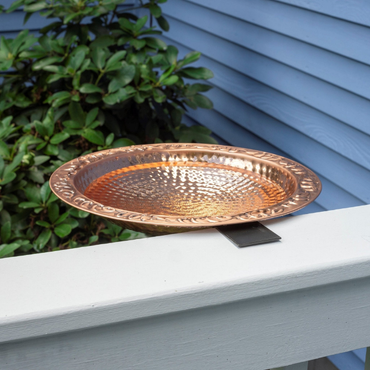 A close-up of the round, hammered copper birdbath attached securely to a black metal deck clamp.