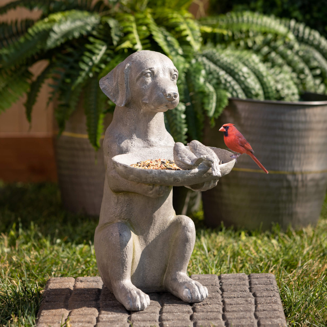 A charming resin dog bird feeder surrounded by lush greenery, with a small bird enjoying the spacious seed dish.