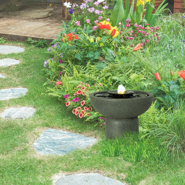 The gray resin water fountain placed among lush greenery and colorful flowers, enhancing outdoor aesthetics.